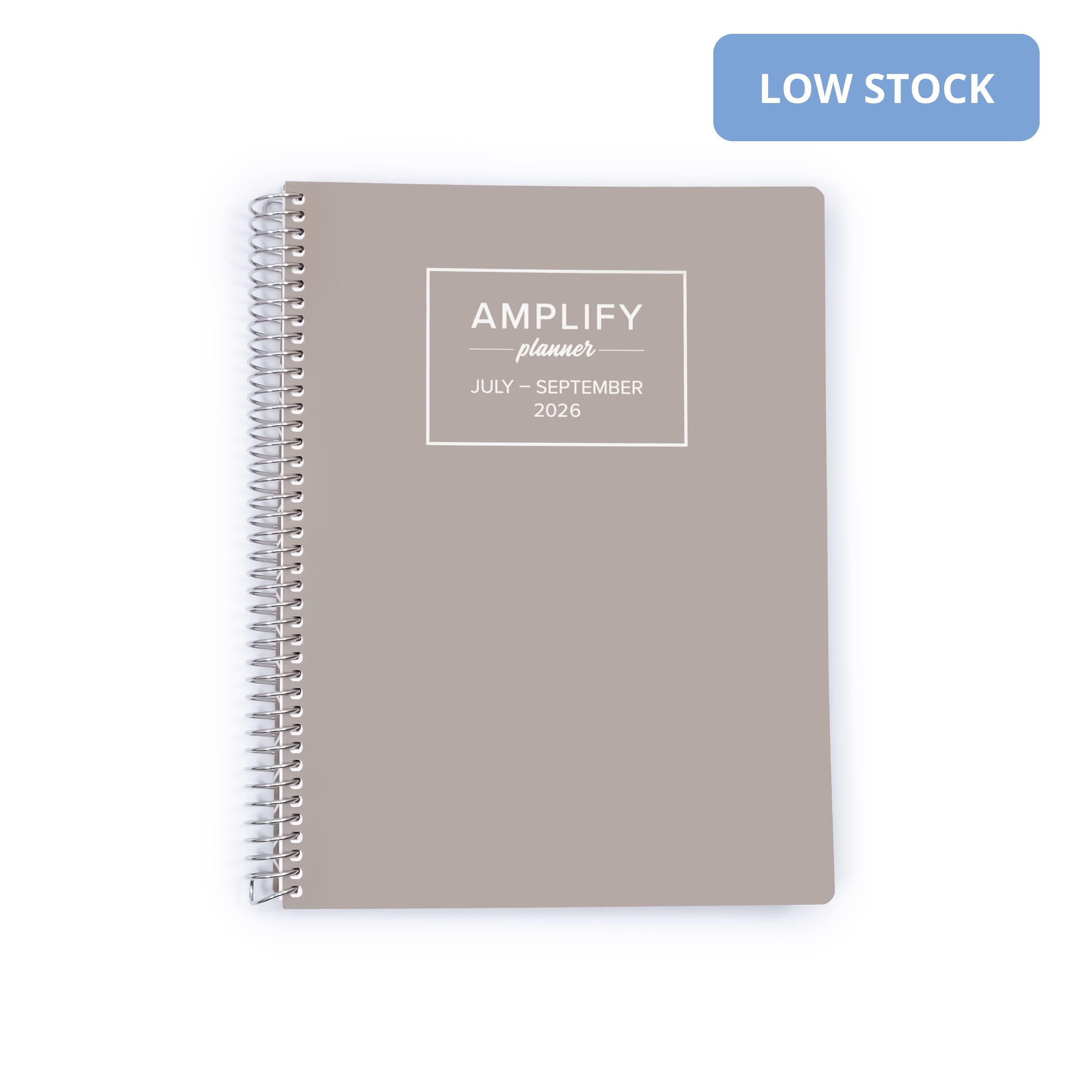 A taupe spiral-bound planner with a white printed label reading 'AMPLIFY planner JULY - SEPTEMBER 2026' on a white background, with a blue 'LOW STOCK' badge in the top right corner.