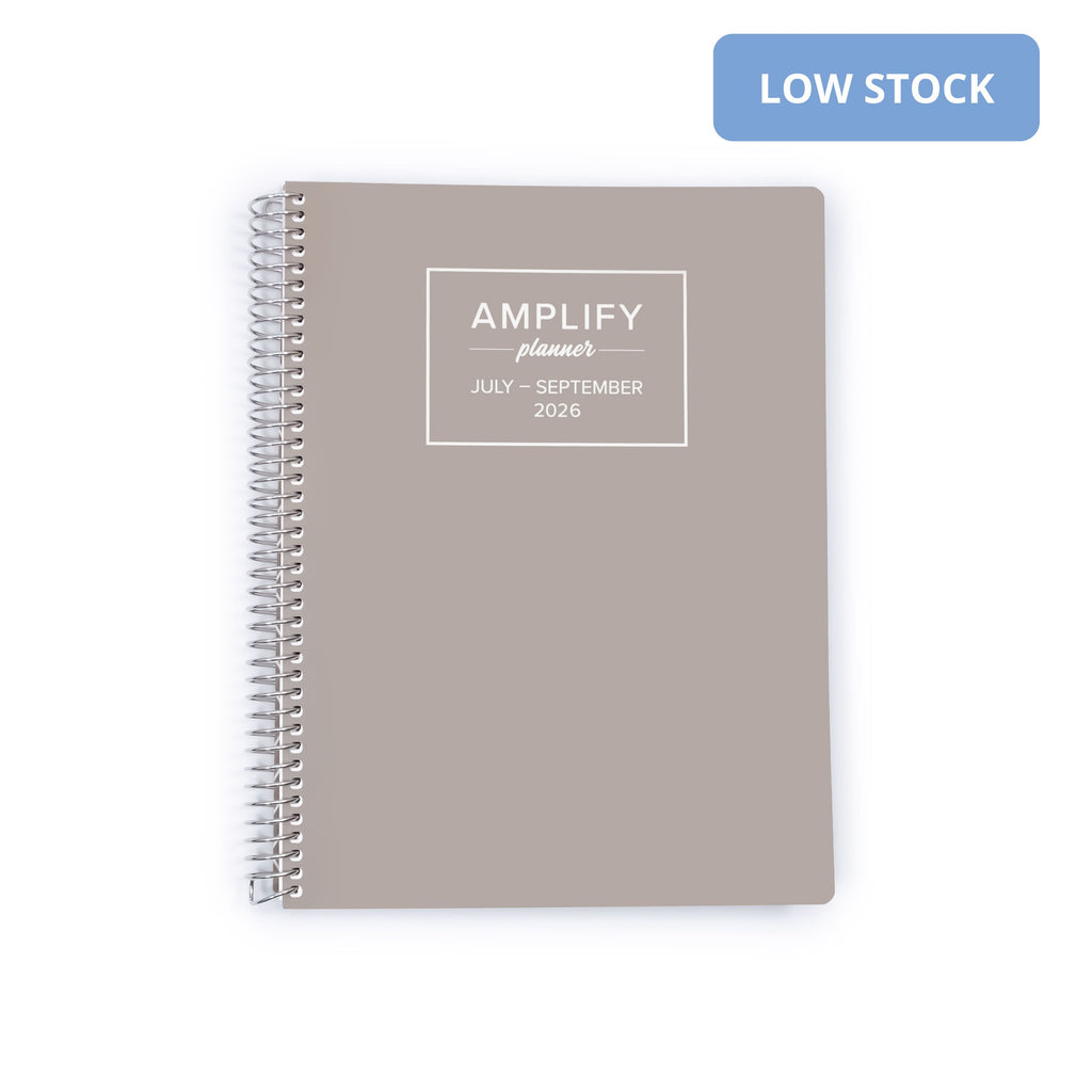 A taupe spiral-bound planner with a white printed label reading 'AMPLIFY planner JULY - SEPTEMBER 2026' on a white background, with a blue 'LOW STOCK' badge in the top right corner.