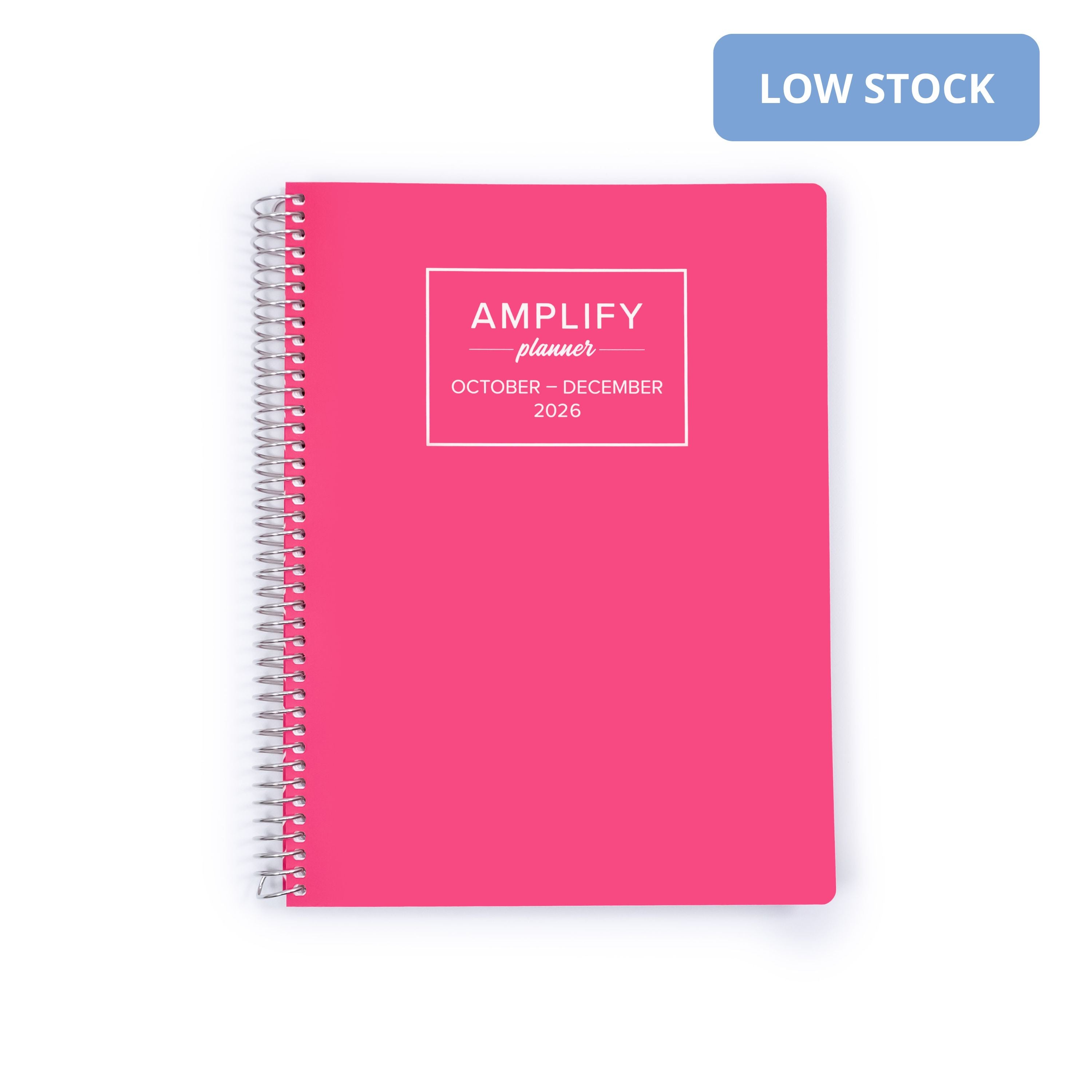 A bright pink spiral-bound planner on a white background with a blue 'LOW STOCK' label in the top right corner. The cover features a white square frame containing the text 'AMPLIFY planner OCTOBER - DECEMBER 2026'.