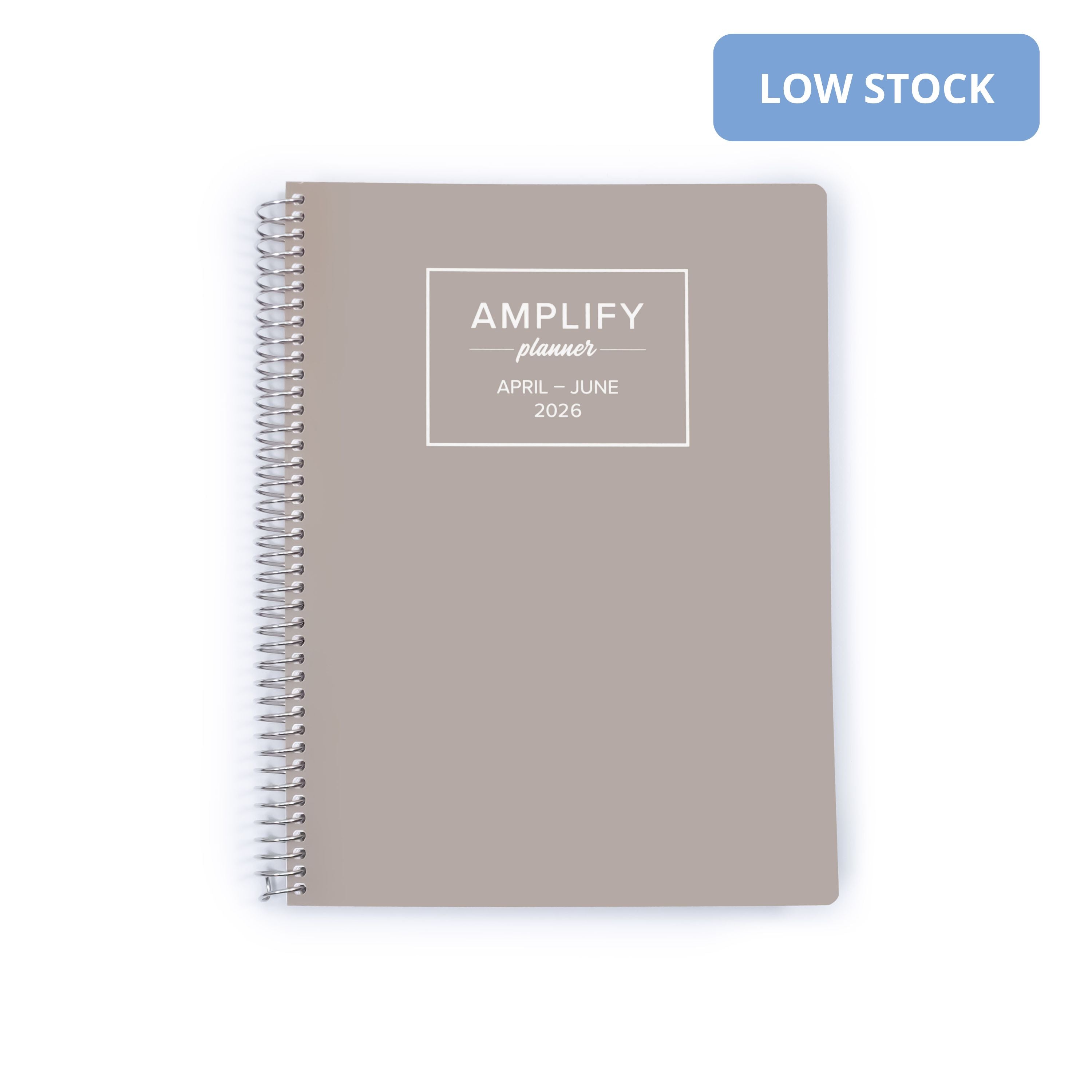 A taupe gray spiral-bound planner on a white background with a blue 'LOW STOCK' badge in the top right corner. The cover features a white square frame containing the text 'AMPLIFY planner APRIL - JUNE 2026'.