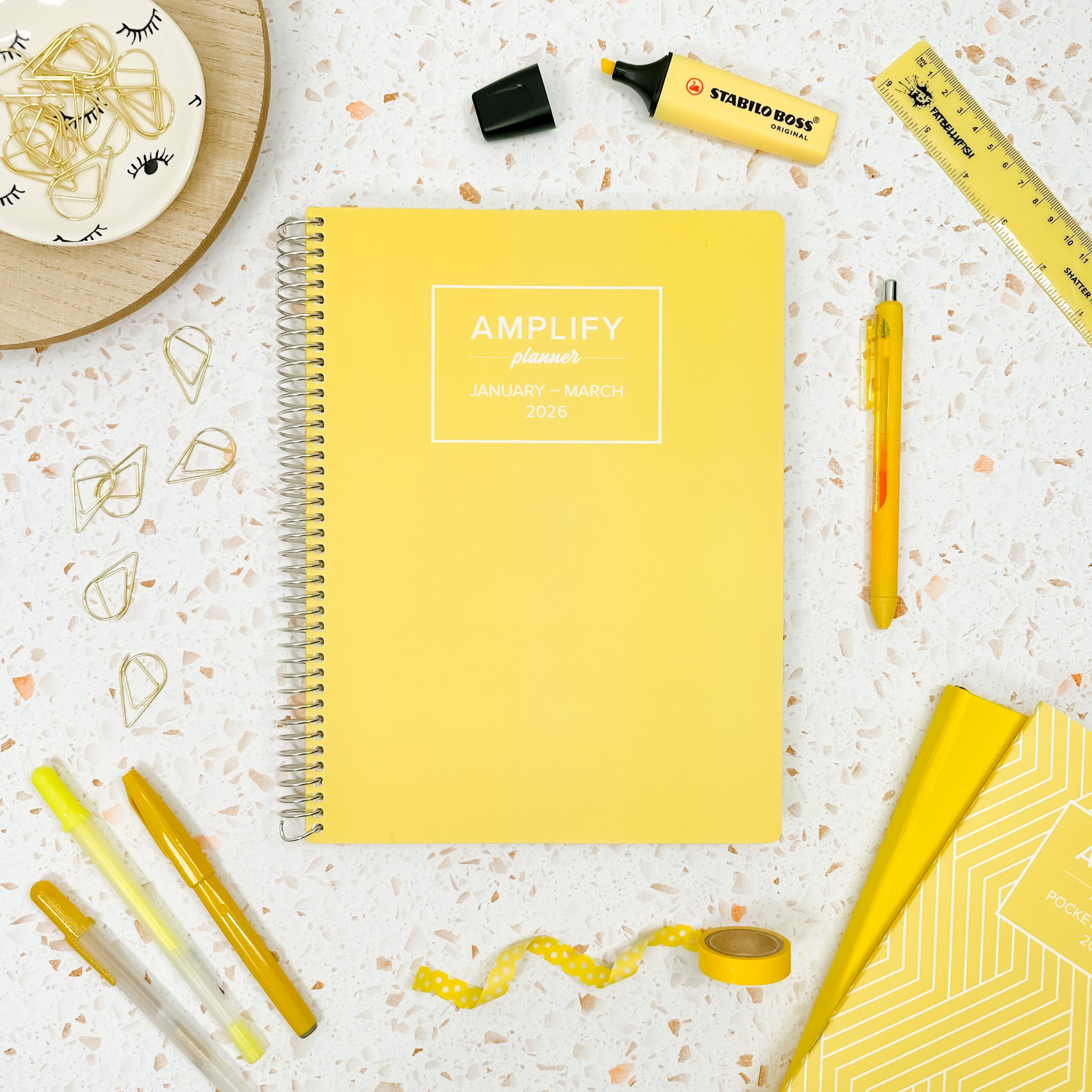 Daily Planner - January-March 2026 - Sunlit Start by Amplify Planner, open on a desk surrounded by office supplies, showcasing its vibrant yellow cover.