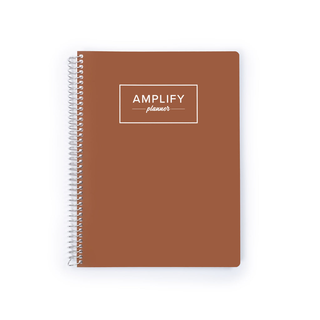 A brown spiral-bound planner with a white wire binding and a white rectangular logo on the front that reads 'AMPLIFY planner', shown on a plain white background.