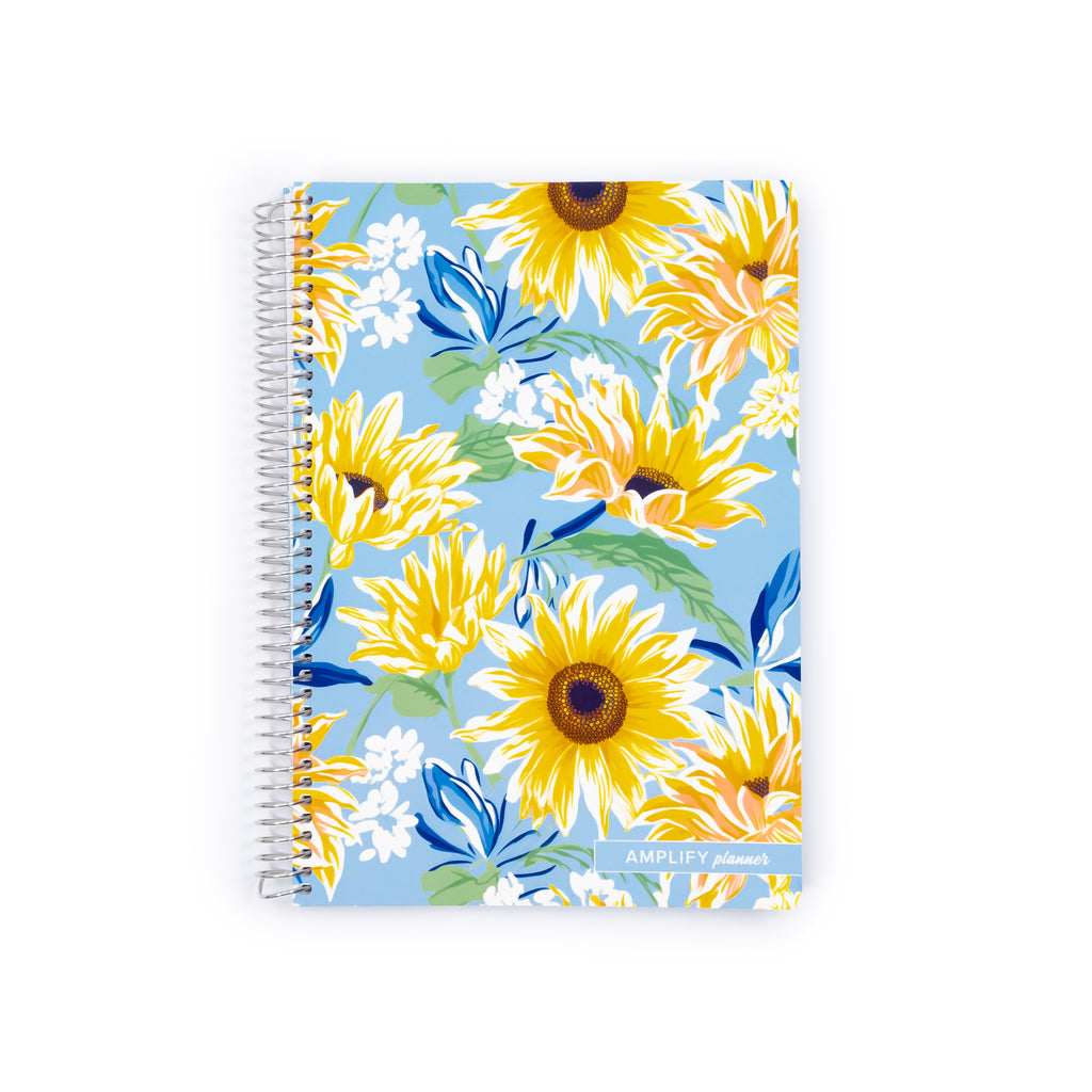 A spiral-bound notebook featuring a vibrant floral pattern of yellow sunflowers, white blossoms, and blue leaves on a light blue background. The brand name 'AMPLIFY stationery' is printed in white on the bottom right corner.
